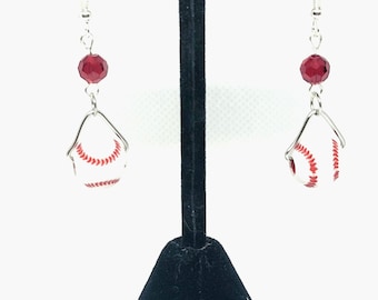 May include: Baseball-themed earrings. Each earring features a miniature baseball design with red stitching, suspended from a silver wire and a faceted red bead. The earrings are displayed on a black stand against a white background.