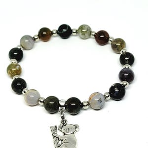 May include: A beaded bracelet featuring a variety of colorful, round gemstones in shades of green, brown, black, and gray. The bracelet has a silver koala charm hanging from the center. The bracelet is on a white background.