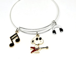 Puede incluir: Pulsera de plata con tres dijes: una nota musical, un perro de dibujos animados tocando la guitarra y unos auriculares.