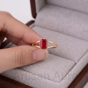 May include: A gold ring featuring a central emerald-cut red gemstone held by four prongs. The band is split into two thin, parallel gold wires that meet at the base of the setting, creating a delicate and refined appearance for the jewelry piece.