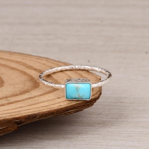 May include: A silver ring featuring a rectangular turquoise stone. The band has a textured, hammered design. The turquoise stone is a bright, vibrant blue-green colour. The ring is displayed on a wooden surface.
