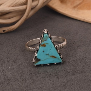 May include: A silver ring featuring a triangular turquoise stone. The turquoise stone has brown veining and is set in a decorative silver bezel. The ring band has a textured design.