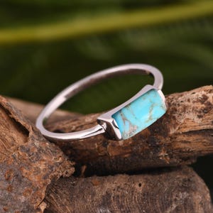 Pode incluir: Um anel de prata com uma pedra turquesa retangular. A pedra azul brilhante tem veios castanhos e está definida num design simples e moderno. O anel é exibido em um pedaço de madeira, com um fundo verde desfocado.
