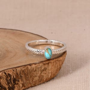 May include: A silver ring with a turquoise stone. The ring has a braided band design and an oval-shaped turquoise stone. The ring is displayed on a wooden surface.