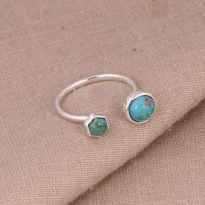 Puede incluir: Un anillo de plata con dos piedras turquesas. Una es redonda y la otra hexagonal. El anillo abierto tiene las piedras en los extremos opuestos de la banda. Las piedras turquesas tienen un aspecto moteado.