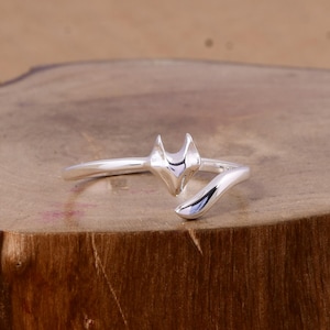 Puede incluir: Anillo plateado con diseño de zorro. El anillo presenta una cabeza y una cola de zorro, con la cola envolviendo el dedo. El anillo es de metal brillante y reflectante, sobre una superficie de madera.