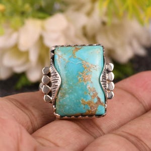 May include: A turquoise ring with a rectangular turquoise stone set in a silver-colored metal band. The turquoise stone has brown veining and is surrounded by a decorative metal frame. The ring has a unique, eye-catching design.