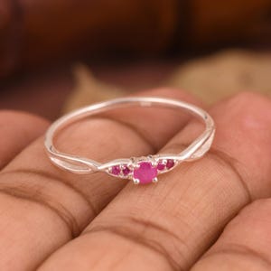 May include: A silver ring with a twisted band design. The ring features a central, round, bright pink gemstone, flanked by smaller pink gemstones. The ring is set against a blurred background.