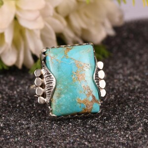 May include: A silver ring featuring a large, rectangular turquoise stone with brown veining. The stone is set in a decorative silver bezel with ornate side details. The ring is displayed on a dark, textured surface, showcasing the vibrant blue-green color of the gemstone.