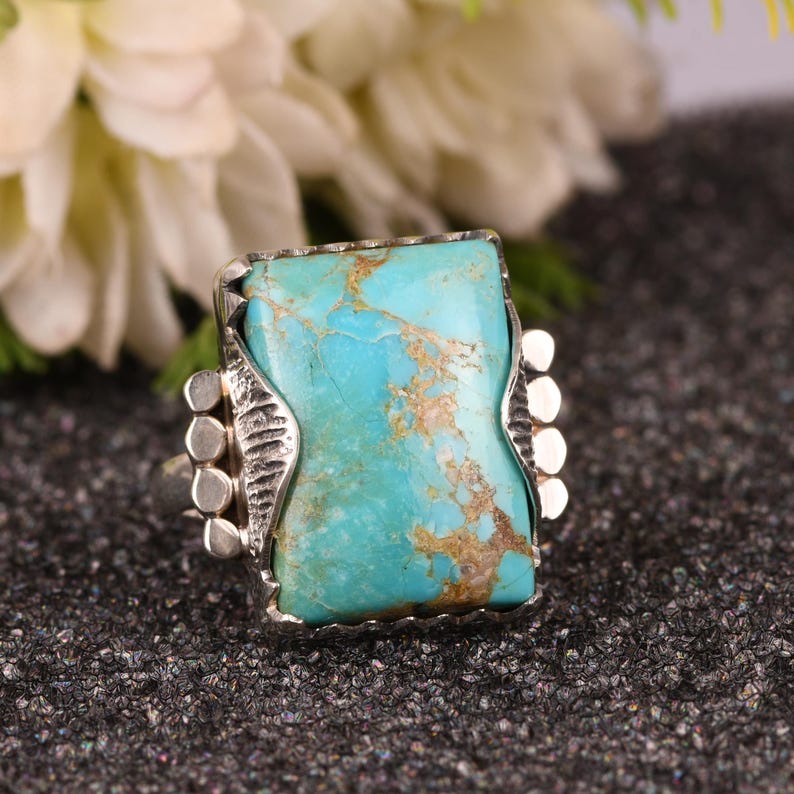 May include: A silver ring featuring a large, rectangular turquoise stone with brown veining. The stone is set in a decorative silver bezel with ornate side details. The ring is displayed on a dark, textured surface, showcasing the vibrant blue-green color of the gemstone.