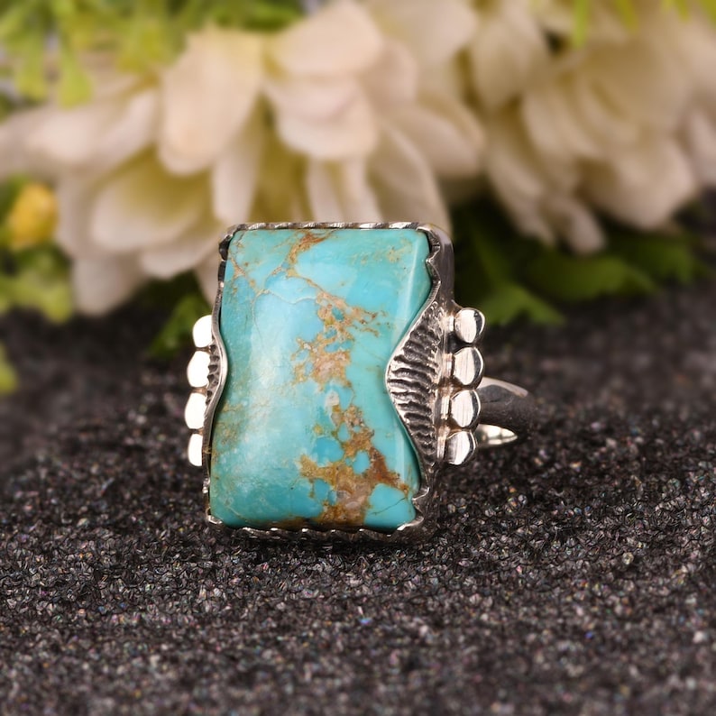 May include: A rectangular turquoise ring with a silver setting. The turquoise stone has brown veining. The ring has decorative silver accents on the sides. The ring is set against a dark, textured background.