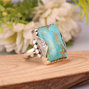 May include: A silver ring featuring a large, rectangular turquoise stone with brown veining. The ring has a decorative silver setting with three small, round accents on the side. The turquoise stone is approximately 1 inch long and 0.75 inches wide.