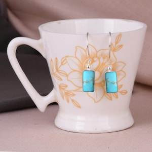 Puede incluir: Un par de pendientes colgantes de plata con piedras turquesas rectangulares que presentan vetas marrones naturales. Los pendientes tienen ganchos finos de plata y cuelgan de una taza de cerámica blanca decorada con un patrón floral de color naranja claro.