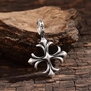Op de afbeelding: Zilveren hanger met een fleur-de-lis ontwerp. De hanger heeft een lus aan de bovenkant met kleine kruisdetails. Het hoofdontwerp is een vierpuntig kruis met gebogen, bloembladachtige elementen. Gepresenteerd op een houten oppervlak.
