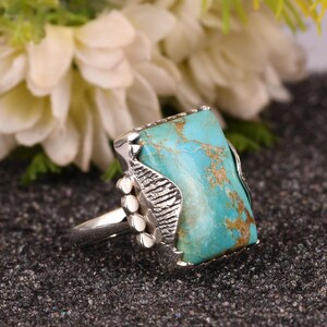 May include: A silver ring featuring a large, rectangular turquoise stone with brown veining. The ring has a detailed, textured setting and three small, round accents on the side. The turquoise stone is a vibrant blue-green color.