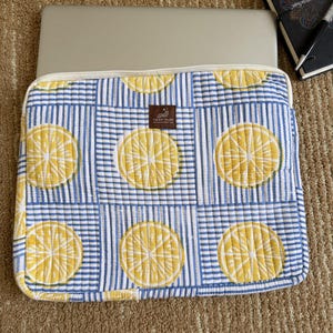 May include: A laptop sleeve with a lemon slice design on a blue and white striped fabric. The sleeve has a cream-coloured border and a label that reads "THE ART RIVER". A laptop is partially visible inside the sleeve.