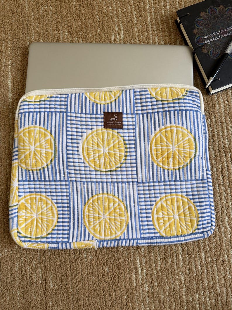 May include: A laptop sleeve with a lemon slice design on a blue and white striped fabric. The sleeve has a cream-coloured border and a label that reads "THE ART RIVER". A laptop is partially visible inside the sleeve.