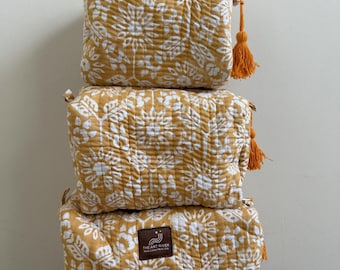 May include: Three stacked, rectangular, quilted cosmetic bags. The bags are mustard yellow with a white floral pattern. Each bag has a matching orange tassel. The bottom bag has a brown leather tag with white text.