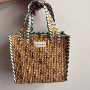 Handmade quilted cotton mini tote bag in mustard yellow with traditional floral block print, green striped handles, and soft padded construction.