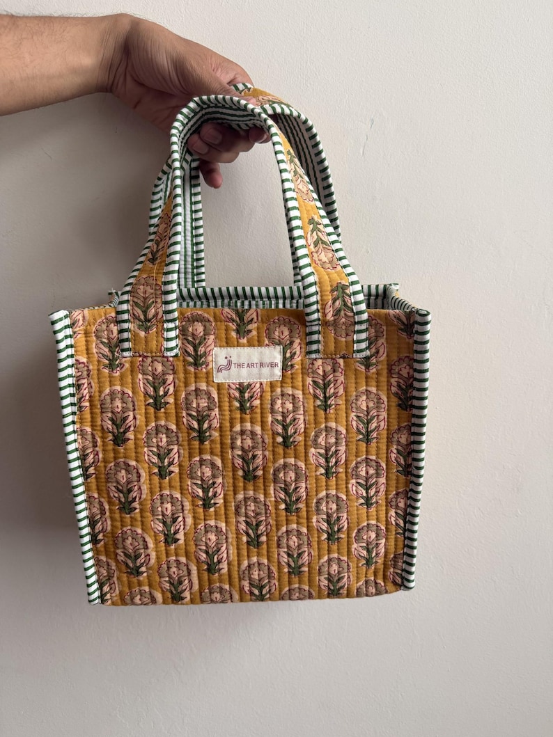 Handmade quilted cotton mini tote bag in mustard yellow with traditional floral block print, green striped handles, and soft padded construction.