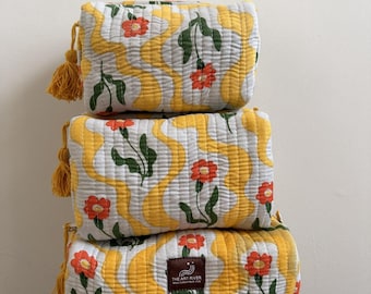 May include: Three stacked, rectangular cosmetic bags with a floral pattern. The bags feature a white background with yellow wavy lines and red flowers. Each bag has a yellow tassel. The bottom bag has a brown label with white text.