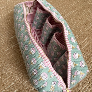 Open quilted cotton makeup organizer bag with four interior brush holder pockets, soft pink striped lining, and spacious zippered compartment for cosmetics and toiletries.