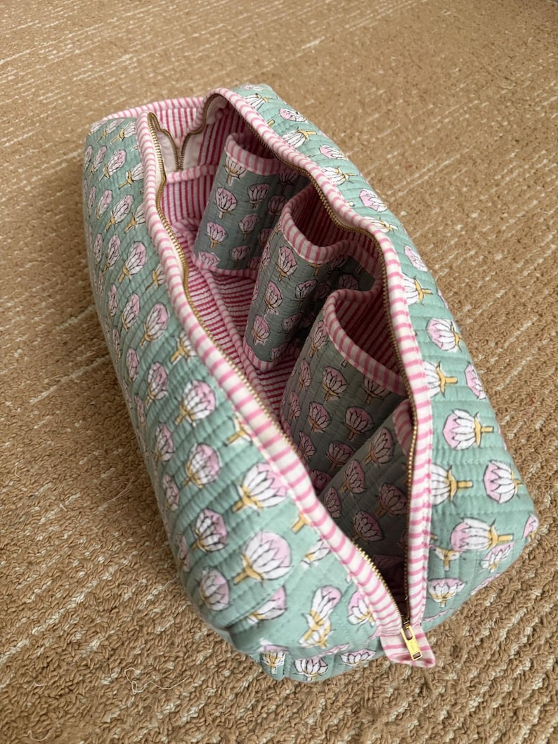 Open quilted cotton makeup organizer bag with four interior brush holder pockets, soft pink striped lining, and spacious zippered compartment for cosmetics and toiletries.