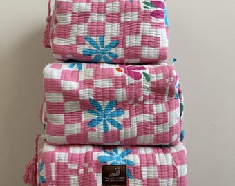 May include: Three stacked, rectangular, quilted cosmetic bags. The bags feature a pink and white checkered pattern with blue and pink floral accents. Each bag has a pink tassel and a label that reads "THE ART RIVER".