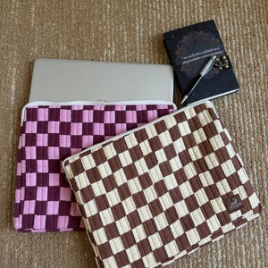 May include: Two laptop cases with a checkered pattern in pink and brown, a notebook and a pen. The pink case has a light and dark pink checkerboard design. The brown case has a brown and cream checkerboard design. The notebook is black with gold accents.