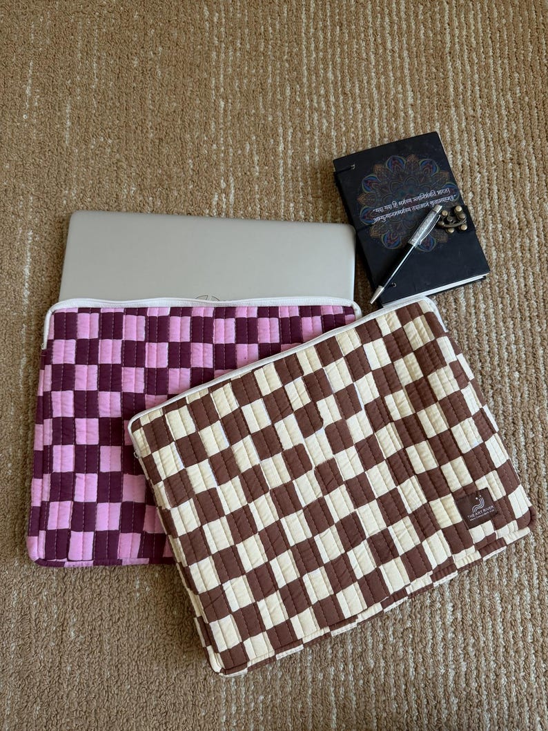 May include: Two laptop cases with a checkered pattern in pink and brown, a notebook and a pen. The pink case has a light and dark pink checkerboard design. The brown case has a brown and cream checkerboard design. The notebook is black with gold accents.
