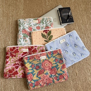 May include: Five rectangular quilted laptop sleeves featuring various floral and striped patterns in colors including pink, blue, green, and orange. Each sleeve has a small white rectangular label with text. A laptop, a black notebook, and a pen are visible.