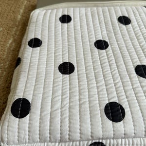 May include: White quilted laptop sleeve with large black polka dots. The sleeve has a rectangular shape with a zip closure and a light grey trim. The fabric is stitched in vertical lines.