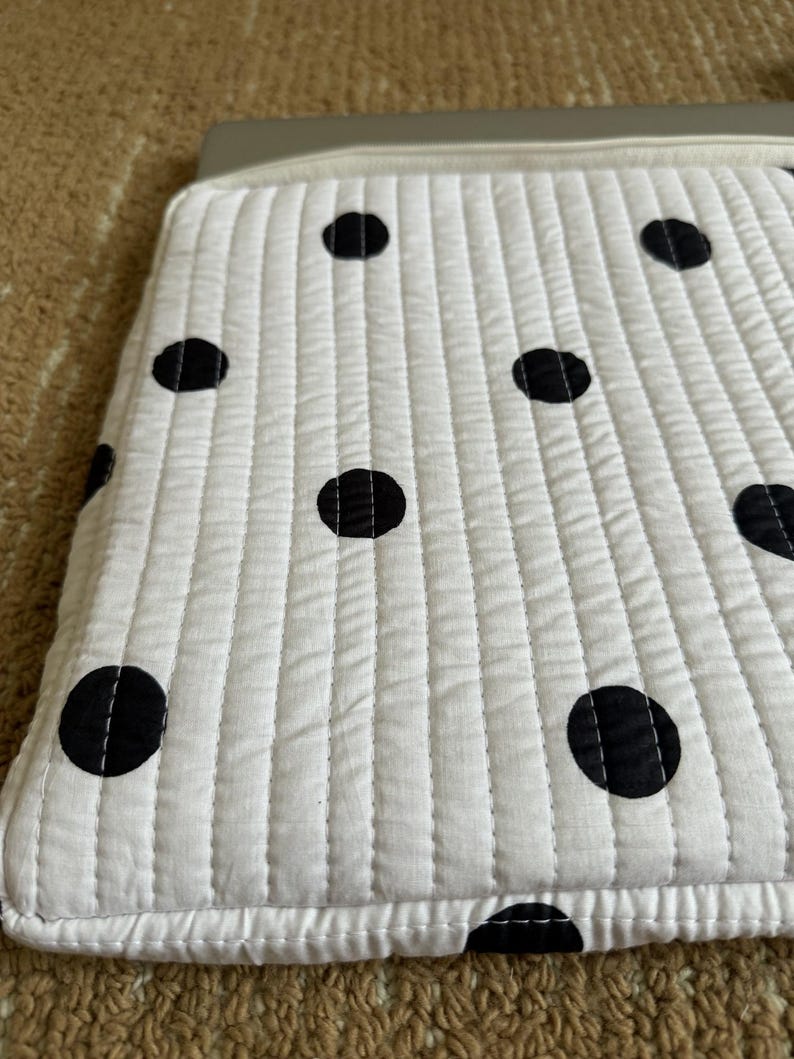 May include: White quilted laptop sleeve with large black polka dots. The sleeve has a rectangular shape with a zip closure and a light grey trim. The fabric is stitched in vertical lines.