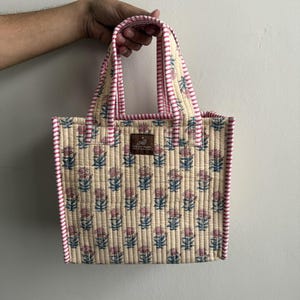 Handmade quilted cotton mini tote bag in ivory cream with delicate blue and pink floral motifs, striped pink handles, and structured silhouette.