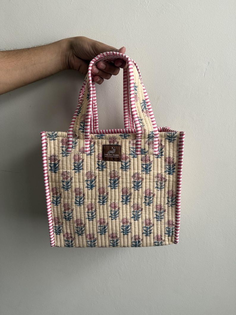 Handmade quilted cotton mini tote bag in ivory cream with delicate blue and pink floral motifs, striped pink handles, and structured silhouette.