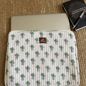May include: White quilted laptop sleeve featuring a repeating pattern of green cacti in pink and blue pots. The sleeve has a green striped border and a brown label. A laptop is inside.
