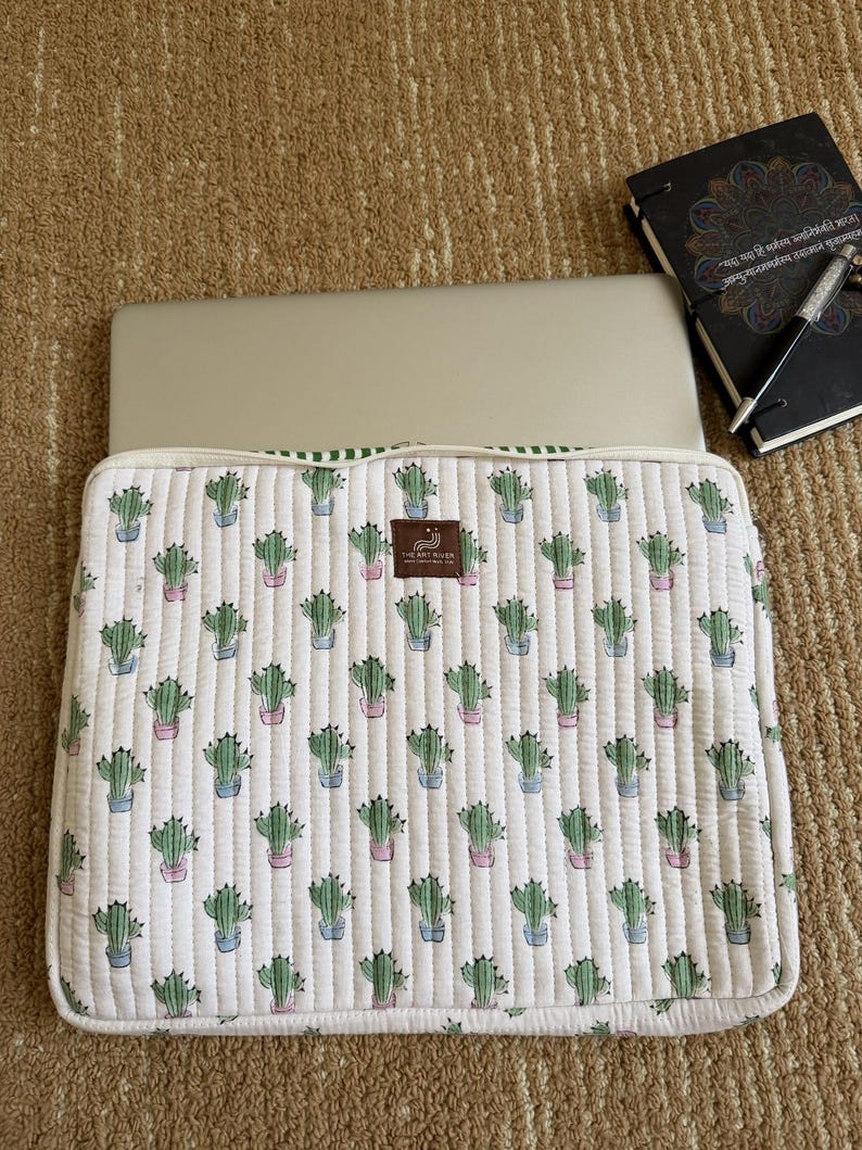 May include: White quilted laptop sleeve featuring a repeating pattern of green cacti in pink and blue pots. The sleeve has a green striped border and a brown label. A laptop is inside.
