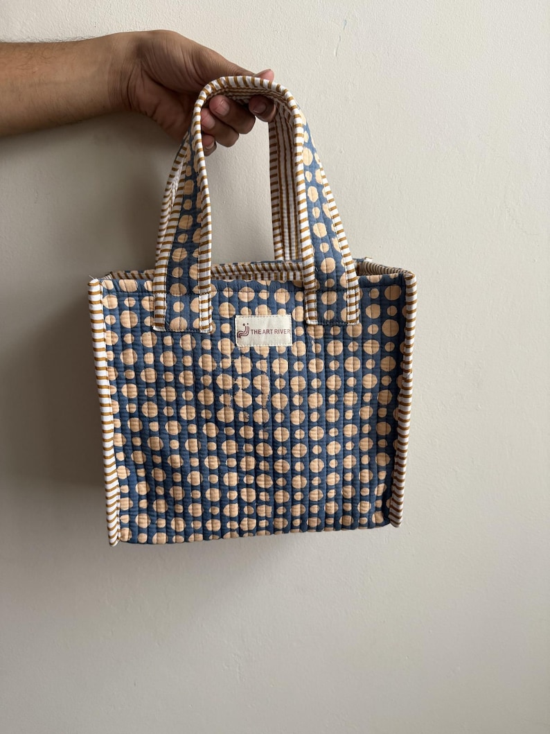 Handmade quilted cotton mini tote bag in indigo blue with beige polka dot print, striped mustard handles, and structured design for carrying daily essentials.