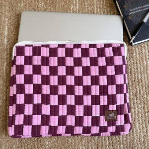 May include: A pink and burgundy checkered laptop sleeve with a white zip. The sleeve is made of quilted fabric and has a small tag with text. A silver laptop is inside the sleeve.