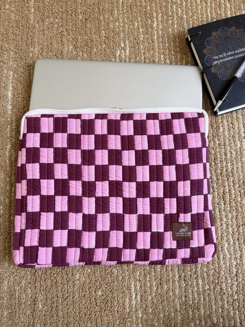May include: A pink and burgundy checkered laptop sleeve with a white zip. The sleeve is made of quilted fabric and has a small tag with text. A silver laptop is inside the sleeve.
