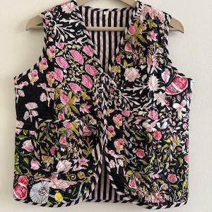 Könnte beinhalten: Eine gesteppte Weste mit schwarzem Hintergrund und einem lebendigen Blumenmuster in Rosa, Weiß und Grün. Die Weste hat einen V-Ausschnitt und ein gestreiftes Futter. Sie hängt an einem Holzbügel.