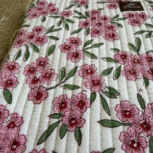 May include: A white quilted laptop sleeve featuring a floral design of pink flowers and green leaves. The sleeve has vertical stitching and a zip closure. The Art River logo is visible.