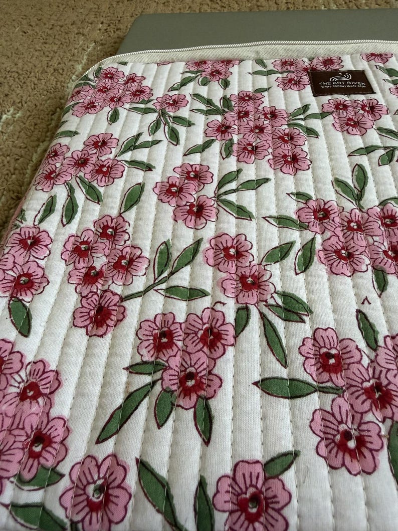 May include: A white quilted laptop sleeve featuring a floral design of pink flowers and green leaves. The sleeve has vertical stitching and a zip closure. The Art River logo is visible.
