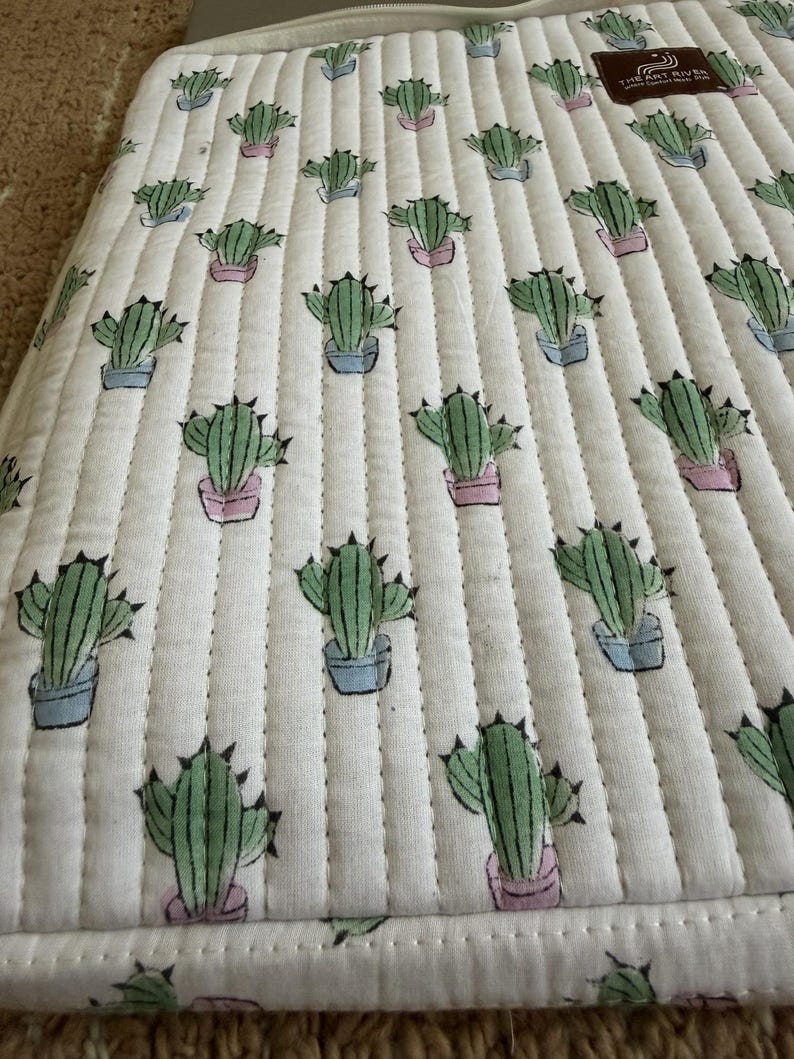 May include: White quilted laptop sleeve featuring a repeating pattern of green cacti in blue and pink pots. The sleeve has a zip closure and a label that reads "The Art River".