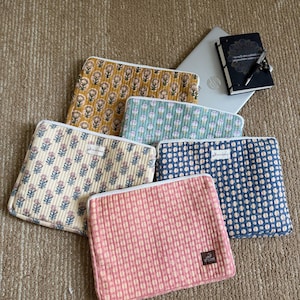 May include: Five rectangular quilted laptop sleeves featuring various patterns including mustard yellow floral, light blue floral, cream floral, blue polka dot, and pink grid. Each sleeve has a white zipper and a small rectangular brand label.