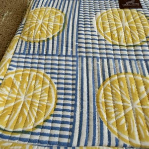 May include: Quilted fabric featuring lemon slice motifs and blue and white stripes. The fabric has a stitched design and a label that reads "THE ART RIVER". The fabric is a light yellow, white, and blue colour.