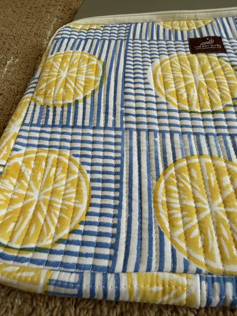 May include: Quilted fabric featuring lemon slice motifs and blue and white stripes. The fabric has a stitched design and a label that reads "THE ART RIVER". The fabric is a light yellow, white, and blue colour.