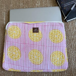 May include: A pink and white striped laptop sleeve featuring a quilted lemon slice design. The sleeve has a white zip and a label reading "THE ART HAVEN". A laptop is inside. A notebook and pen are in the background.