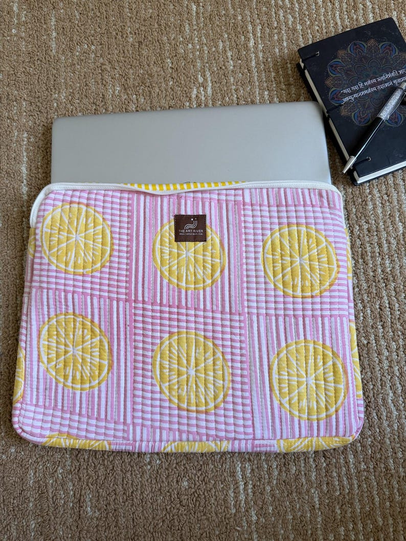 May include: A pink and white striped laptop sleeve featuring a quilted lemon slice design. The sleeve has a white zip and a label reading "THE ART HAVEN". A laptop is inside. A notebook and pen are in the background.