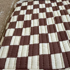 May include: Close-up of a quilted fabric with a brown and cream checkerboard pattern. The fabric is stitched with a linear pattern. The edge of the fabric is visible, with a grey border.
